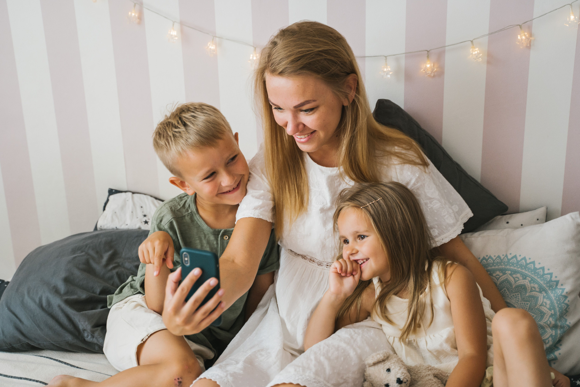 A mom in a sunlit kitchen, smiling at her phone — kids' artwork pinned to the fridge behind her